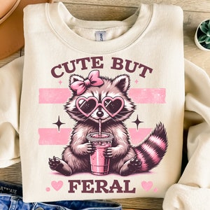 Op de afbeelding: Crèmekleurige sweatshirt met een illustratie van een wasbeer met een hartvormige zonnebril en een strik. De wasbeer drinkt een roze drankje. De tekst "CUTE BUT FERAL" staat boven en onder de wasbeer.