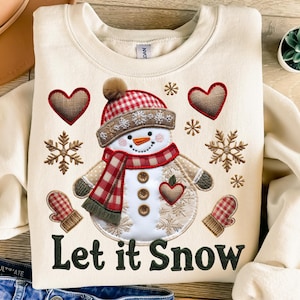 Peut inclure: Sweat-shirt crème avec un motif de bonhomme de neige. Le bonhomme de neige porte un chapeau et une écharpe à carreaux rouges et blancs, avec des flocons de neige, des cœurs et des moufles brodés. Le texte "Let it Snow" est imprimé en vert foncé.