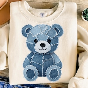 May include: A cream-coloured sweatshirt with a denim patchwork teddy bear design. The bear is made from various shades of blue denim, with black eyes and a black nose. The sweatshirt has a crew neck and long sleeves.