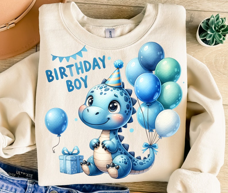 May include: A light beige sweatshirt with a blue dinosaur wearing a party hat and holding blue balloons. The text "Birthday Boy" is printed on the sweatshirt.