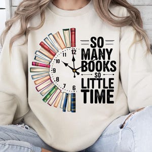 K&ouml;nnte beinhalten: Ein wei&szlig;es Sweatshirt mit einer Grafik eines Zifferblatts, das von gestapelten B&uuml;chern umgeben ist. Der Text lautet "So Many Books So Little Time".
