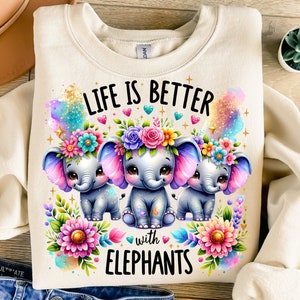 May include: A cream-coloured sweatshirt with the text "Life is better with elephants" in black. The text is surrounded by colourful flowers and three cartoon elephants wearing flower crowns.