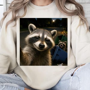 May include: Cream-colored sweatshirt featuring a square graphic of two raccoons. One raccoon is taking a selfie, while the other peeks out from behind a blue trash bin. The image is set at night with streetlights in the background.