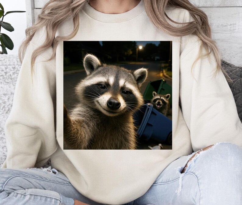 May include: Cream-colored sweatshirt featuring a square graphic of two raccoons. One raccoon is taking a selfie, while the other peeks out from behind a blue trash bin. The image is set at night with streetlights in the background.