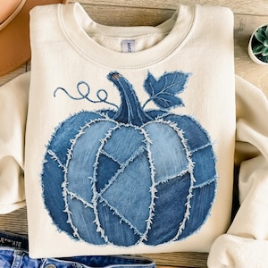 May include: Cream-colored sweatshirt featuring a large blue denim pumpkin design. The pumpkin is made of various denim patches with visible stitching, a stem, and a leaf. The design is centered on the front of the long-sleeved top.