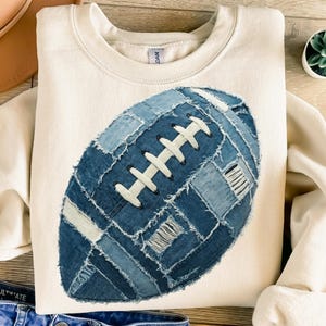 May include: Cream-colored sweatshirt featuring a large, denim patchwork football design. The football is made of various shades of blue denim with white stitching details. A casual, sporty apparel item.