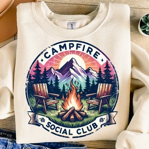 May include: Cream-colored sweatshirt with a circular graphic featuring a campfire scene. The design includes a mountain range, trees, a bonfire, two wooden chairs, and the text "CAMPFIRE SOCIAL CLUB". The graphic uses shades of blue, pink, orange, and green.