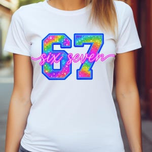 K&ouml;nnte beinhalten: Wei&szlig;es T-Shirt mit einem farbenfrohen, regenbogenfarbenen "67"-Design, umrandet in Blau. Die Worte "six seven" sind in einer kursiven, rosa Schrift &uuml;ber den Zahlen geschrieben.