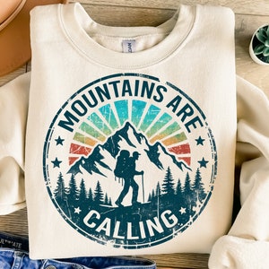 Puede incluir: Una sudadera beige con un estampado gráfico vintage de una cordillera con un excursionista y el texto "Mountains are calling".