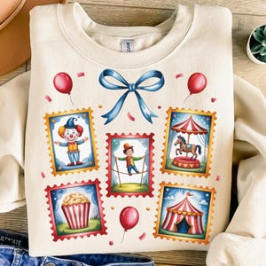 May include: Cream-colored sweatshirt featuring a whimsical circus-themed design. The design includes illustrations of a clown, a tightrope walker, a carousel horse, popcorn, and a circus tent, all framed as postage stamps. Red balloons and a blue bow complete the design.