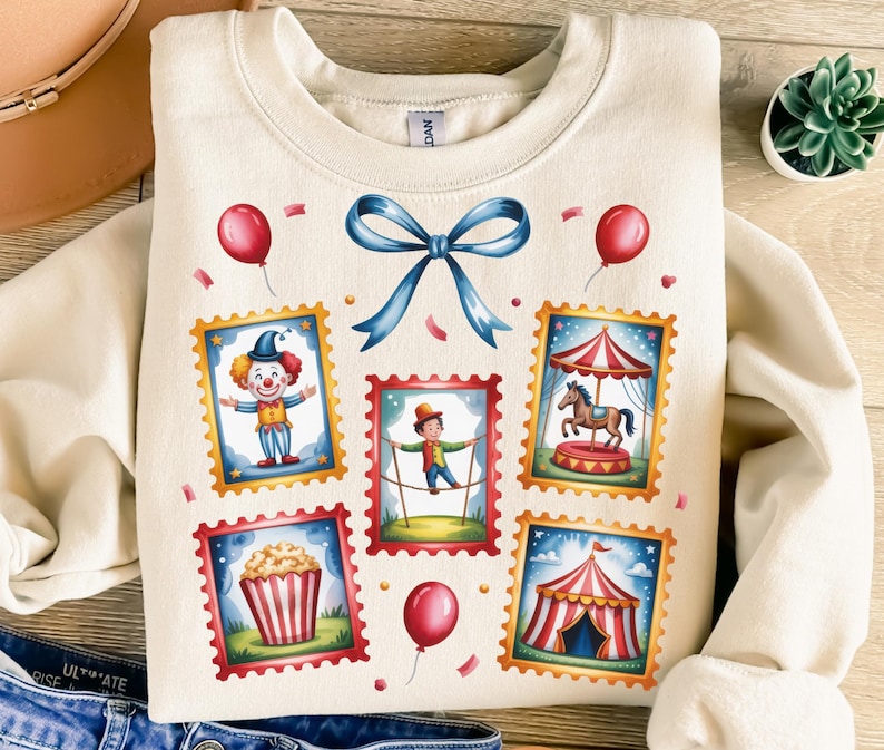 May include: Cream-colored sweatshirt featuring a whimsical circus-themed design. The design includes illustrations of a clown, a tightrope walker, a carousel horse, popcorn, and a circus tent, all framed as postage stamps. Red balloons and a blue bow complete the design.