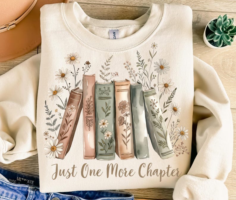 Op de afbeelding: Cr&egrave;mekleurige sweatshirt met een bloemen- en boekontwerp. De afbeelding bevat madeliefjes, groen en boeken in aquarel. De tekst "Just One More Chapter" is gedrukt in een bruin script. De sweatshirt heeft een ronde hals.