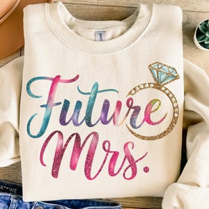 May include: A cream-coloured sweatshirt with the text "Future Mrs." in a rainbow-coloured font with glitter accents. A gold glitter ring with a blue diamond is above the text.