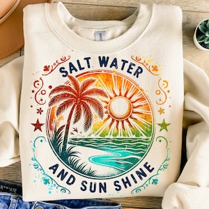 May include: A cream-colored sweatshirt with a vintage-style graphic print of a palm tree, sun, and ocean waves. The text "Salt Water and Sun Shine" is printed in a circle around the graphic.