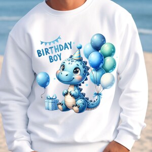 May include: White sweatshirt with a blue dinosaur wearing a party hat and holding balloons. The text "Birthday Boy" is printed on the shirt.