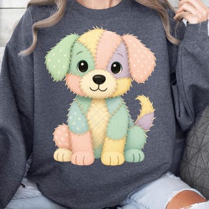 May include: A dark grey sweatshirt with a colourful patchwork puppy design. The puppy has large eyes and is made up of pastel-coloured fabric patches, including pink, yellow, green, and blue. The design is centred on the front of the sweatshirt.