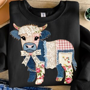Op de afbeelding: Zwart sweatshirt met een patchwork koeienontwerp. De koe heeft denim, kant en bloemenaccenten, met een roze en wit geruit patroon op haar lichaam. Een witte kanten strik siert de nek en de hoorns zijn versierd met roze rozen.