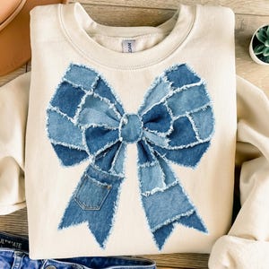 May include: Cream-colored sweatshirt featuring a large bow design made from various shades of blue denim patches. The bow has a pocket detail on one side. The sweatshirt is a casual, comfortable style.