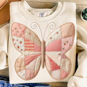 May include: Cream-coloured sweatshirt with a patchwork butterfly design in pink, beige, and white. The butterfly wings are made of various fabric patterns, including gingham and floral prints. The sweatshirt has a crew neck and long sleeves.
