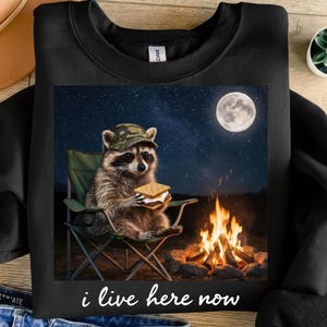 Puede incluir: Sudadera negra con un mapache con sombrero de camuflaje, sosteniendo un s'more y sentado en una silla de camping. La imagen incluye una fogata, una luna llena y el texto "i live here now".