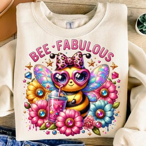 May include: Cream-colored sweatshirt with a colorful graphic of a bee wearing heart-shaped glasses and a bow. The bee is surrounded by flowers and a drink, with the words "BEE FABULOUS" above. The design is vibrant and whimsical.