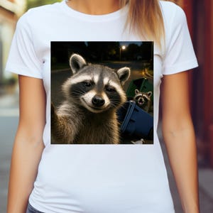 May include: White t-shirt featuring a square image of a raccoon taking a selfie near a blue trash bin. The raccoon has a gray and black mask and is smiling. Another raccoon is visible in the background.