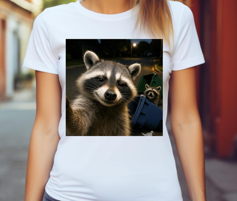 May include: White t-shirt featuring a square image of a raccoon taking a selfie near a blue trash bin. The raccoon has a gray and black mask and is smiling. Another raccoon is visible in the background.