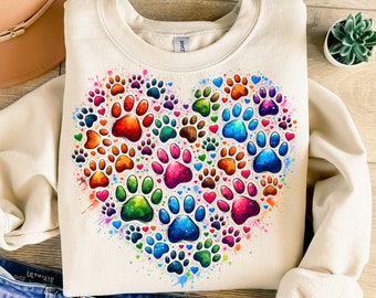 May include: A cream-colored sweatshirt with a colorful heart design made of paw prints. The paw prints are in shades of red, orange, yellow, green, blue, and purple. The heart is surrounded by smaller paw prints and hearts.