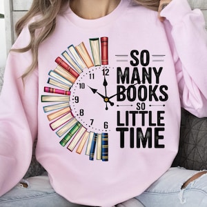 K&ouml;nnte beinhalten: Rosafarbener Pullover mit einem Grafikdruck einer Uhr aus B&uuml;chern. Der Text auf dem Pullover lautet "So many books, so little time."