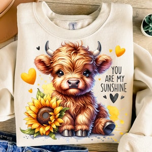 May include: A beige sweatshirt with a cute cartoon Highland cow wearing a sunflower crown. The text "You are my sunshine" is printed on the sweatshirt.