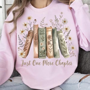 Op de afbeelding: Een lichtroze sweatshirt met een aquarelontwerp van boeken en bloemen. Het ontwerp bevat madeliefjes en andere bloemelementen rond een rij boeken. De tekst "Just One More Chapter" is in goud gedrukt.