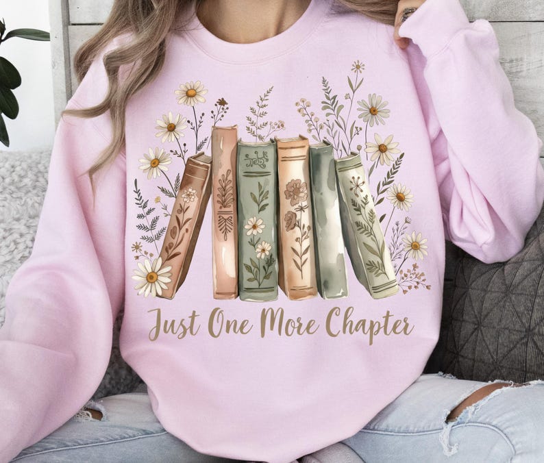 Op de afbeelding: Een lichtroze sweatshirt met een aquarelontwerp van boeken en bloemen. Het ontwerp bevat madeliefjes en andere bloemelementen rond een rij boeken. De tekst "Just One More Chapter" is in goud gedrukt.