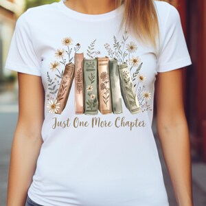 Op de afbeelding: Wit T-shirt met een aquarel illustratie van boeken en bloemen. Het ontwerp bevat de tekst "Just One More Chapter" in goud. De boeken zijn in tinten bruin, groen en roze, met madeliefjes en andere bloemen.