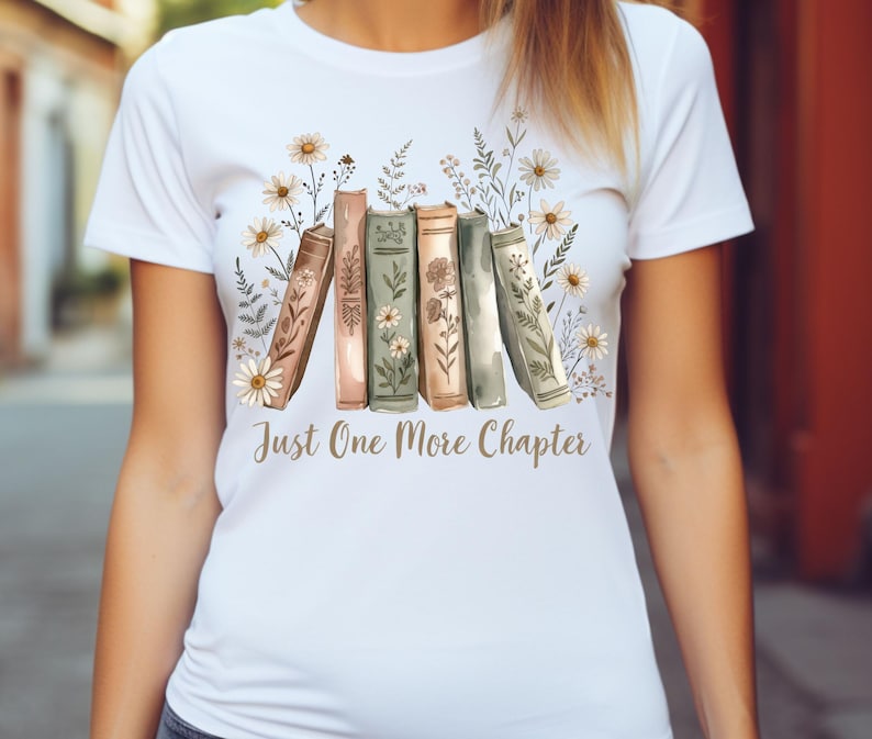 Op de afbeelding: Wit T-shirt met een aquarel illustratie van boeken en bloemen. Het ontwerp bevat de tekst "Just One More Chapter" in goud. De boeken zijn in tinten bruin, groen en roze, met madeliefjes en andere bloemen.