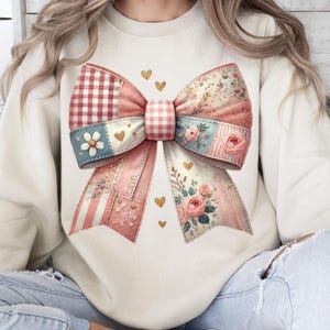 May include: Cream-colored sweatshirt featuring a large, patchwork bow design. The bow incorporates various patterns, including gingham, floral, and striped elements, in shades of pink, red, and blue. Gold heart accents are scattered around the bow.