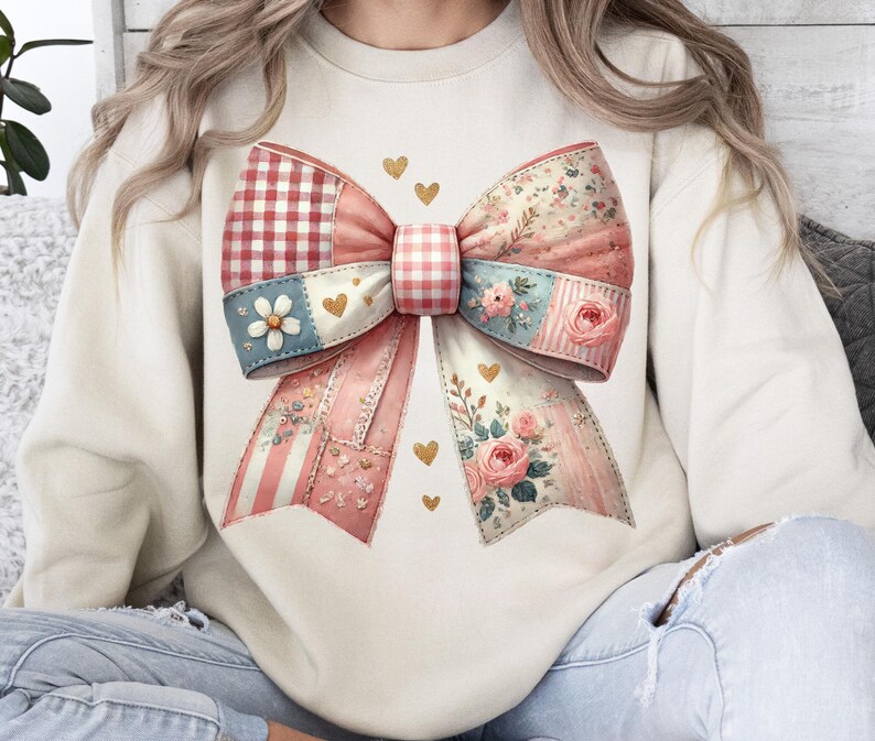 May include: Cream-colored sweatshirt featuring a large, patchwork bow design. The bow incorporates various patterns, including gingham, floral, and striped elements, in shades of pink, red, and blue. Gold heart accents are scattered around the bow.