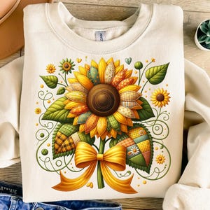 Pode incluir: Camisola creme com um design detalhado de girassol. O girassol tem um padrão de retalhos em amarelo, laranja, verde e castanho. Um laço amarelo está na base da flor. Elementos florais adicionais e folhas verdes rodeiam o girassol.