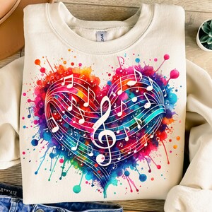 Pode incluir: Uma camisa de moletom branca com um design de coração de aquarela colorido com notas musicais e uma clave de sol. O coração é cercado por respingos de tinta coloridos.