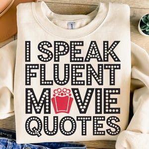May include: A cream-colored sweatshirt with the text "I Speak Fluent Movie Quotes" in black letters with white dots around each letter. The text is in a marquee style font.