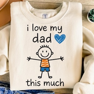 Può includere: Felpa color crema con la scritta "i love my dad this much" in nero, un cuore blu e un'illustrazione di un omino stilizzato. L'omino ha una maglietta arancione e pantaloncini blu. È visibile una piccola pianta succulenta.