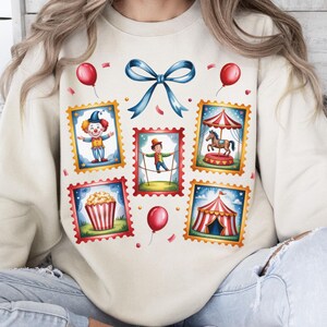 May include: Cream-colored sweatshirt featuring a whimsical circus-themed design. The design includes postage stamp-style illustrations of a clown, tightrope walker, carousel, popcorn, and a circus tent, along with red balloons and a blue bow.