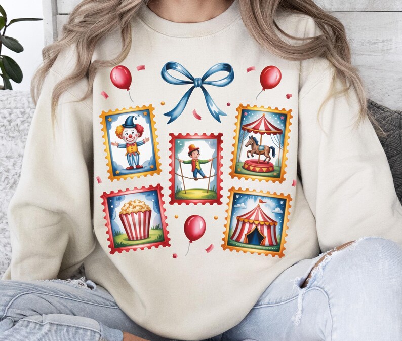 May include: Cream-colored sweatshirt featuring a whimsical circus-themed design. The design includes postage stamp-style illustrations of a clown, tightrope walker, carousel, popcorn, and a circus tent, along with red balloons and a blue bow.