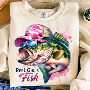 May include: A beige sweatshirt featuring a graphic of a bass fish wearing pink sunglasses and a pink camouflage hat. The fish is surrounded by pink watercolour splatters. The text "Reel Girls Fish" is printed below the fish.