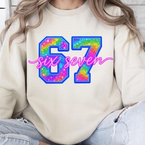 K&ouml;nnte beinhalten: Cremefarbenes Sweatshirt mit einem gro&szlig;en, farbenfrohen "67"-Design. Die Zahlen sind blau umrandet und mit einem Regenbogen-Glitzer-Muster gef&uuml;llt. Die Worte "six seven" sind in einem rosa Skript &uuml;ber den Zahlen geschrieben.