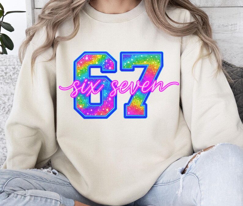 K&ouml;nnte beinhalten: Cremefarbenes Sweatshirt mit einem gro&szlig;en, farbenfrohen "67"-Design. Die Zahlen sind blau umrandet und mit einem Regenbogen-Glitzer-Muster gef&uuml;llt. Die Worte "six seven" sind in einem rosa Skript &uuml;ber den Zahlen geschrieben.