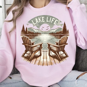 May include: Light pink sweatshirt featuring a lakeside graphic. The design includes two wooden Adirondack chairs on a dock, facing a lake with mountains, trees, and the text "LAKE LIFE" in a banner.
