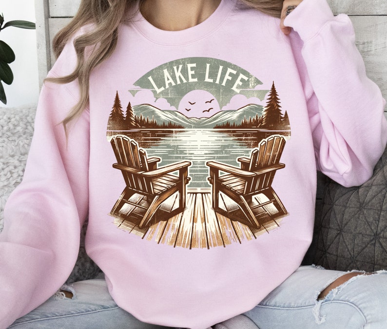 May include: Light pink sweatshirt featuring a lakeside graphic. The design includes two wooden Adirondack chairs on a dock, facing a lake with mountains, trees, and the text "LAKE LIFE" in a banner.
