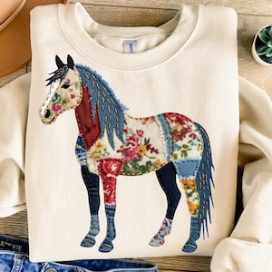 May include: Cream-colored sweatshirt featuring a patchwork horse design. The horse has a floral pattern on its body, with blue, red, and floral accents. The horse's mane and tail are blue. The sweatshirt is a casual, comfortable style.
