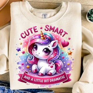 Könnte beinhalten: Cremefarbenes Sweatshirt mit einem bunten Einhorn-Motiv. Das Einhorn hat ein goldenes Horn, eine rosa und lila Mähne und ist von Sternen und Herzen umgeben. Der Text lautet "CUTE + SMART AND A LITTLE BIT DRAMATIC."