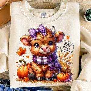 Op de afbeelding: Crèmekleurige sweatshirt met een cartoon Highland-koe met een paarse geruite strik en sjaal, omringd door pompoenen. De koe staat tegen een achtergrond van herfstbladeren en de tekst "Hay Fall". Een charmant herfstthema-ontwerp.